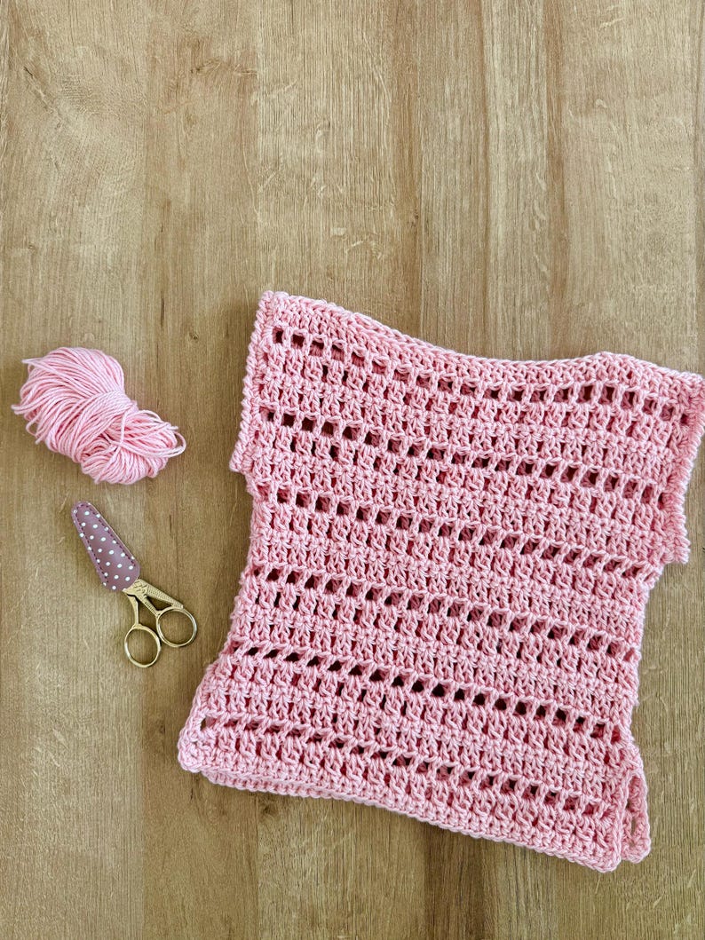 May include: A light pink crochet top featuring a repeating openwork stitch pattern, accompanied by a small ball of matching pink yarn and a pair of gold-toned embroidery scissors with a brown and white polka-dot fabric cover on the blades.
