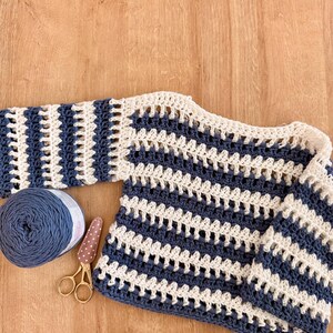 Op de afbeelding: Een gehaakte trui met lange mouwen en horizontale strepen in marineblauw en cr&egrave;me. Het kledingstuk is gemaakt met een open, luchtig steekpatroon. Naast de trui liggen een bol marineblauw garen en een kleine gouden schaar.