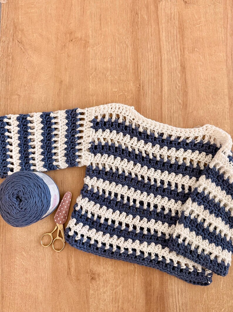 Op de afbeelding: Een gehaakte trui met lange mouwen en horizontale strepen in marineblauw en cr&egrave;me. Het kledingstuk is gemaakt met een open, luchtig steekpatroon. Naast de trui liggen een bol marineblauw garen en een kleine gouden schaar.