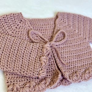 Pu&ograve; includere: Un cardigan all'uncinetto rosa antico con maniche corte e chiusura con fiocco. I bordi del cardigan sono adornati da un motivo floreale smerlato. La maglia morbida e strutturata &egrave; adatta a neonati e bambini piccoli.