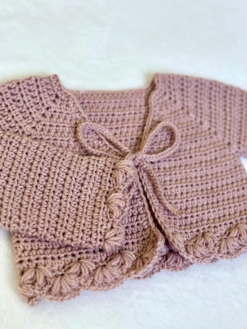 Pu&ograve; includere: Un cardigan all'uncinetto rosa antico con maniche corte e chiusura con fiocco. I bordi del cardigan sono adornati da un motivo floreale smerlato. La maglia morbida e strutturata &egrave; adatta a neonati e bambini piccoli.