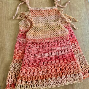 May include: Two sleeveless dresses crafted from crocheted yarn featuring an ombre color gradient that transitions from light peach at the top to a deep coral red at the hem. Each dress has thin shoulder straps that tie into bows at the top.