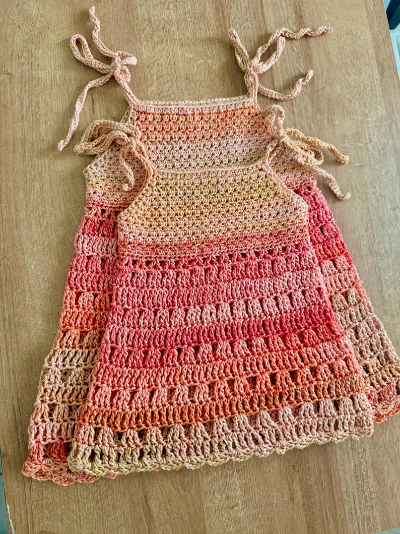 May include: Two sleeveless dresses crafted from crocheted yarn featuring an ombre color gradient that transitions from light peach at the top to a deep coral red at the hem. Each dress has thin shoulder straps that tie into bows at the top.