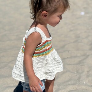 May include: A white crochet top with a colorful striped design across the chest. The top has a square neckline and wide straps. The bottom of the top flares out. The top is paired with denim shorts.