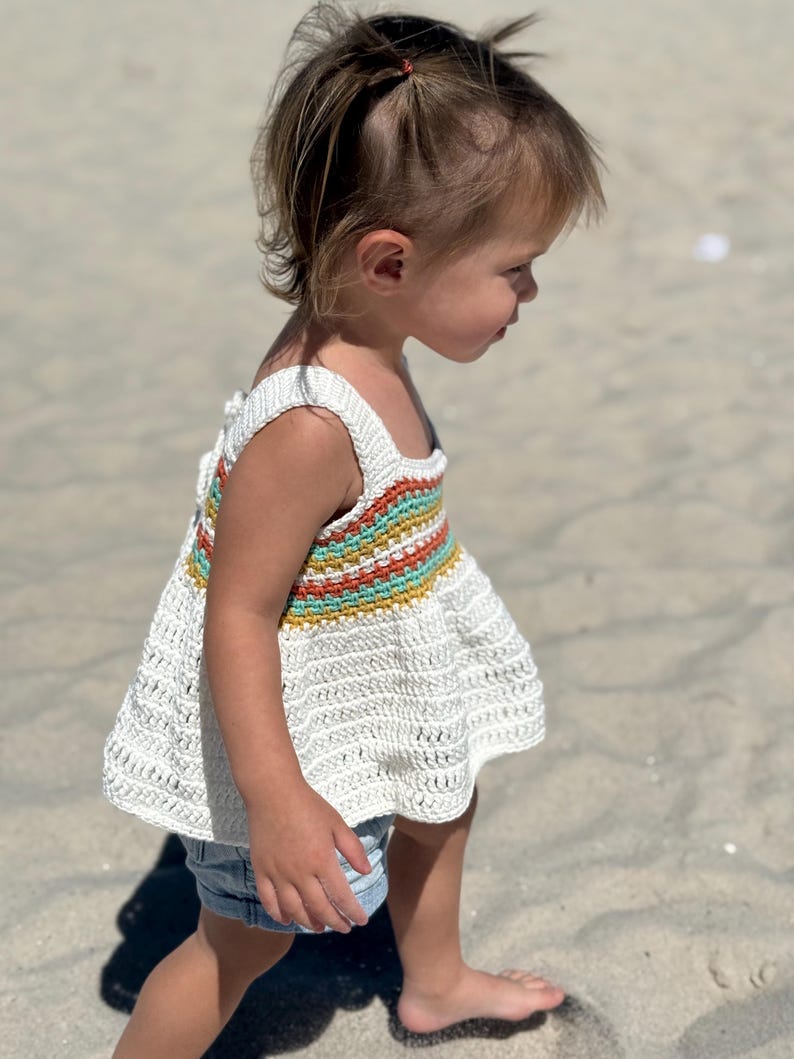 May include: A white crochet top with a colorful striped design across the chest. The top has a square neckline and wide straps. The bottom of the top flares out. The top is paired with denim shorts.
