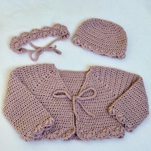 Pu&ograve; includere: Un set all'uncinetto in tre pezzi di colore rosa antico. Il set comprende un cardigan con chiusura a fiocco, una fascia e un cappello a cuffia. I bordi del cardigan e del cappello presentano un design a conchiglia. Il set &egrave; probabilmente per un bambino o un bambino piccolo.