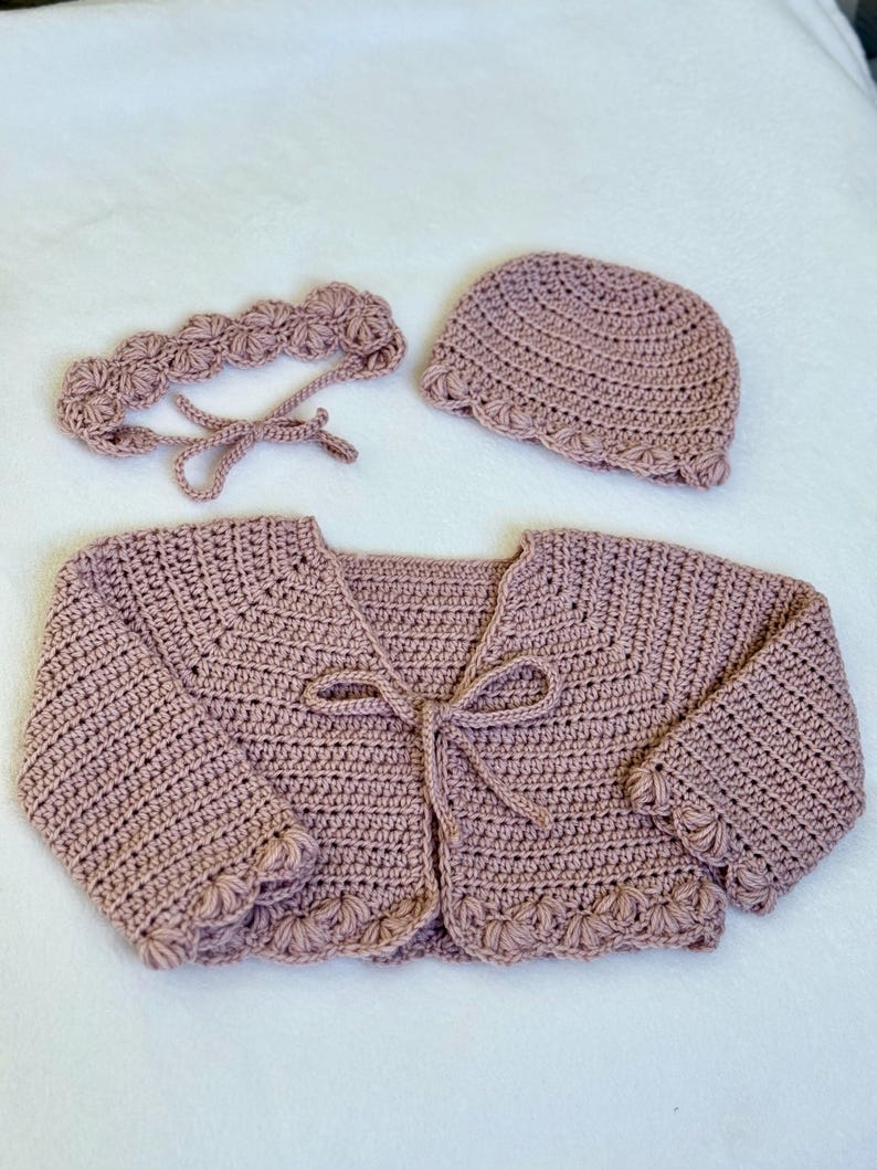 Pu&ograve; includere: Un set all'uncinetto in tre pezzi di colore rosa antico. Il set comprende un cardigan con chiusura a fiocco, una fascia e un cappello a cuffia. I bordi del cardigan e del cappello presentano un design a conchiglia. Il set &egrave; probabilmente per un bambino o un bambino piccolo.