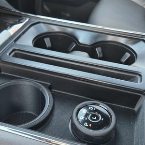 May include: A black car console with two cup holders, a small storage area, and a rotary gear selector. The console is made of black plastic and has a silver trim. The gear selector has the letters "P, R, N, D, L" on it.