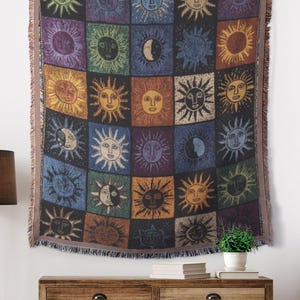 May include: A tapestry featuring a grid of colorful sun and moon designs hangs above a wooden dresser. The tapestry has a fringed edge. A floor lamp with a brown shade and gold legs stands to the left. A woven basket and decorative plants are in front of the dresser.
