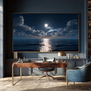 May include: A large framed print depicts a nighttime ocean scene with a full moon reflecting on the water. The artwork hangs above a wooden desk with a brass lamp and a blue armchair in a room with dark blue walls.