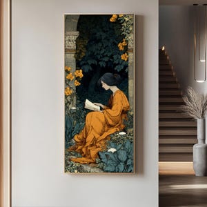 May include: A vertical framed wall art print depicting a person with dark hair wearing a long orange dress, sitting and reading a book within a stone archway surrounded by dark green foliage, blue-tinted plants, and clusters of bright yellow flowers.