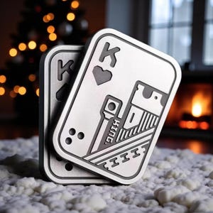 Könnte beinhalten: Zwei silberne Untersetzer in Form von Spielkarten, jeweils mit dem König-Herz-Design. Die Untersetzer haben abgerundete Ecken und detaillierte Gravuren, einschließlich des Buchstabens "K" und des Wortes "King". Der Hintergrund zeigt einen verschwommenen Weihnachtsbaum und einen Kamin.