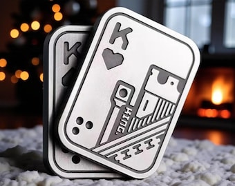 May include: Two silver metal playing card coasters, each featuring the King of Hearts design. The coasters have rounded corners and detailed engravings, including the letter "K" and the word "King". The background shows a blurred Christmas tree and a fireplace.