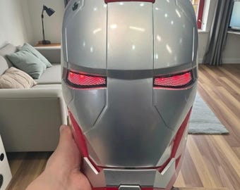 May include: Silver and red Iron Man helmet with glowing red eye slits. The helmet has a sleek, metallic finish and is held by a person. The design is based on the iconic superhero's mask, perfect for cosplay or display.