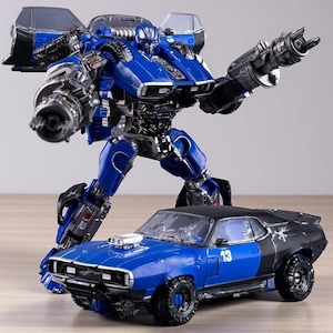 Puede incluir: Una figura de robot de juguete azul y negro con un coche azul y negro a juego. El robot tiene armas grandes y está posado como en acción. El coche tiene el número 13 en el lateral.