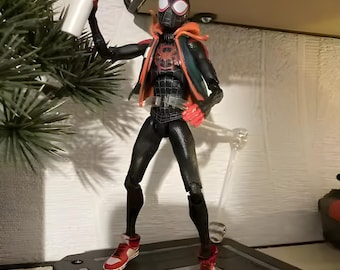 May include: A Miles Morales action figure wearing a black suit with a red spider emblem, an orange hooded jacket, and red and white sneakers. The figure holds a small white spray paint can in one hand and is supported by a clear plastic stand.
