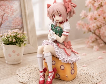 May include: A collectible anime-style figurine of a character with short pink hair and red eyes, wearing a white and pink ruffled dress with bows, white socks, and red shoes. The figure sits on a yellow textured base, holding a green plaid pillow.