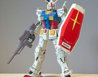 May include: A detailed model of a Gundam robot, primarily white with red, blue, and yellow accents. It holds a gray rifle and a red shield with a yellow star. The robot has intricate mechanical details and is posed upright.