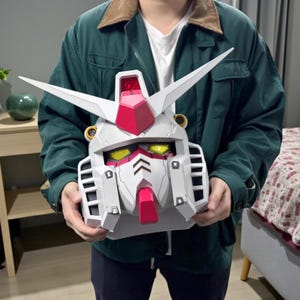 Puede incluir: Una cabeza de robot Gundam blanca, roja y gris con ojos amarillos y dos antenas blancas en forma de V. La cabeza del robot tiene una barbilla roja y una cresta roja en la parte superior. La cabeza es sostenida por una persona.