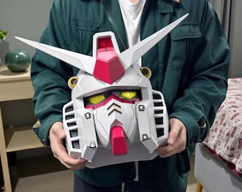 May include: A white, red, and gray Gundam robot head with yellow eyes and two white V-shaped antennas. The robot head has a red chin and a red crest on top. The head is held by a person.