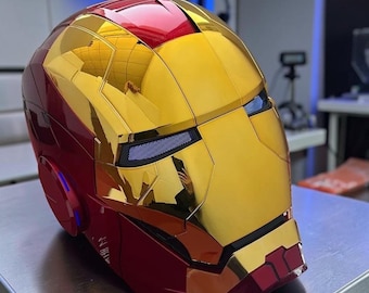May include: A full-face helmet featuring a metallic gold and deep red finish. The design includes a reflective gold faceplate with integrated eye slits containing blue light-emitting mesh panels and circular red side accents on the exterior shell.