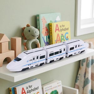May include: A white toy train with blue detailing and "CRH" text is on a white shelf. The train is made up of several connected carriages. Children's books and a green dinosaur rattle are in the background.