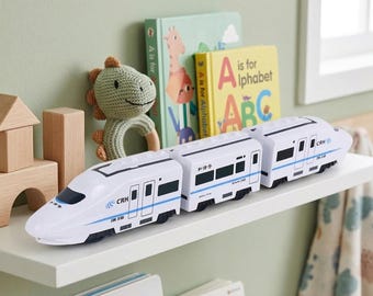 May include: A white toy train with blue accents and the text "CRH" is displayed on a white shelf. The train is composed of multiple connected cars. Children's books and a green dinosaur rattle are in the background.