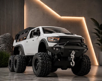 May include: A white and black toy truck with six large black tires. The truck has a black front bumper with a silver hook and a black roof rack. The headlights are orange. The truck is on a gray surface.