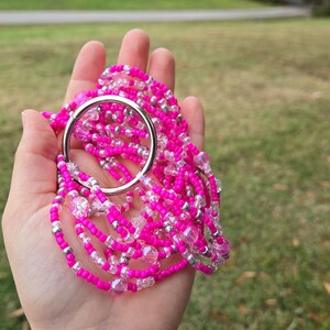 May include: A collection of bright pink beaded strands, accented with clear and silver beads, held together by a large silver ring. The beads are a vibrant fuchsia color, creating a striking contrast against the silver.