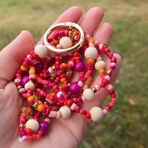 May include: A beaded lanyard with a gold-colored clasp, held in a hand. The lanyard features a mix of red, pink, yellow, and purple beads, along with wooden beads. The background is a blurred view of green grass and trees.