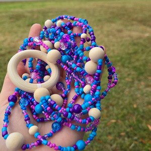 May include: A colorful beaded necklace with a wooden ring and wooden beads. The necklace features blue, purple, and pink beads. The wooden ring is approximately 2 inches in diameter. The necklace is held in a hand against a blurred green background.