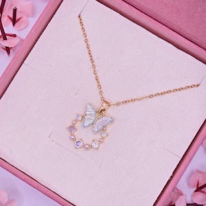 May include: A gold-colored necklace with a butterfly pendant, presented in a pink gift box. The butterfly has light blue and white wings, surrounded by a circle of small, colorful gems. The necklace is delicate and elegant.