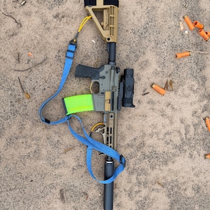 K&ouml;nnte beinhalten: Ein olivgr&uuml;nes und beigefarbenes Gewehr mit einem schwarzen Zielfernrohr, Schalld&auml;mpfer und einem neongr&uuml;nen Magazin. Ein blauer Gurt ist angebracht. Verbrauchte orangefarbene Patronenh&uuml;lsen liegen auf einer sandigen Oberfl&auml;che verstreut.