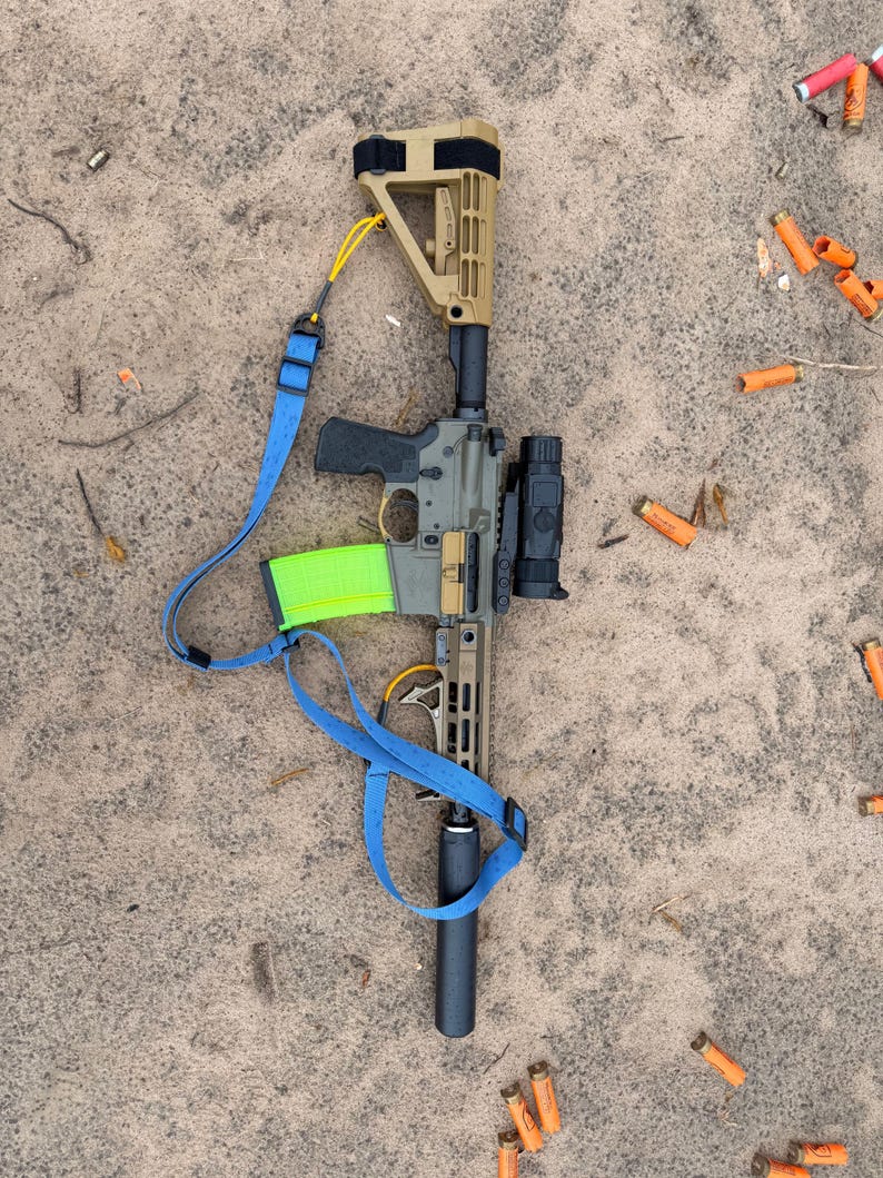 K&ouml;nnte beinhalten: Ein olivgr&uuml;nes und beigefarbenes Gewehr mit einem schwarzen Zielfernrohr, Schalld&auml;mpfer und einem neongr&uuml;nen Magazin. Ein blauer Gurt ist angebracht. Verbrauchte orangefarbene Patronenh&uuml;lsen liegen auf einer sandigen Oberfl&auml;che verstreut.