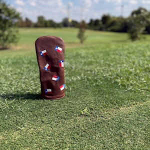 May include: A brown golf club head cover featuring multiple Texas state flags. The cover is made of a soft material and is standing upright on a green grassy field under a blue sky.