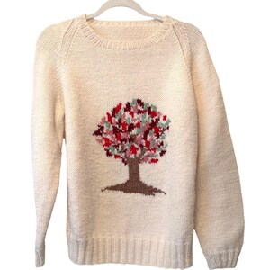 Puede incluir: Suéter de punto color crema con cuello redondo y mangas largas. La parte delantera presenta un tronco de árbol marrón y una colorida copa de árbol con hojas rojas, rosas y verdes. El suéter tiene dobladillo y puños acanalados.