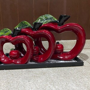 May include: A decorative sculpture featuring three stylized red apple shapes with open centres, accented with green leaves and small red apple details. The sculpture sits on a black rectangular base, suitable for home decor.