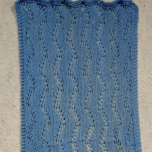 May include: A light blue knitted fabric with a lacy pattern. The fabric is pinned to a white wall.
