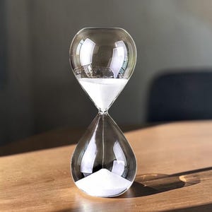 May include: A clear glass hourglass with white sand inside. The hourglass has a bulbous top and bottom, connected by a narrow neck. The sand is flowing from the top to the bottom. The hourglass is sitting on a wooden surface.