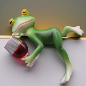 May include: A whimsical green frog figurine with yellow eyes and a red-lipped smile, holding a glass of red wine. The frog is leaning, with one leg extended. The wine glass is clear, and the wine inside is a deep red.