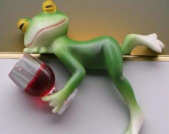 Puede incluir: Una caprichosa figura de rana verde con ojos amarillos y boca roja, sosteniendo una copa de vino tinto. La rana está reclinada, con una pata extendida. La copa de vino es transparente y el vino es rojo oscuro.