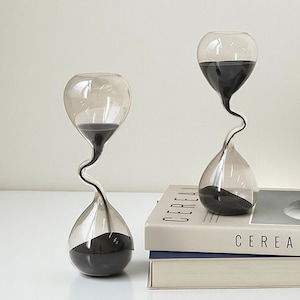 May include: Two clear glass hourglasses with a distinctive, curved design. Each hourglass contains black sand, with the sand in one hourglass partially run through. The hourglasses are placed on a white surface next to a stack of books.