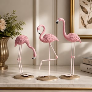 Könnte beinhalten: Ein Set aus drei rosafarbenen Flamingo-Figuren mit strukturierten Körpern, langen dünnen Beinen und schwarz gespitzten Schnäbeln. Jeder Vogel steht auf einem runden goldfarbenen Sockel, wobei ein Flamingo den Hals beugt und einer auf einem Bein steht.