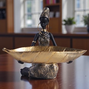 May include: A black and gold figurine of a woman holding a large, leaf-shaped bowl. The woman wears a headdress, hoop earrings, and jewellery. The bowl is gold, and the base has leaf patterns. The figurine is on a wooden surface.