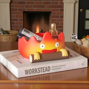 May include: A whimsical, flame-shaped storage container in shades of red, orange, and yellow, with a cartoonish face and arms holding a log. The container sits atop a book with the title "WORKSTEAD".