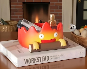 May include: A whimsical, flame-shaped storage container in shades of red, orange, and yellow, with a cartoonish face and arms holding a log. The container sits atop a book with the title "WORKSTEAD".