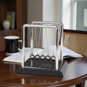 May include: A Newton's cradle, a desktop toy with five polished steel balls suspended from a chrome frame. The black base sits on a dark wooden table. A black mug and open book are also on the table. The table is likely made of wood.