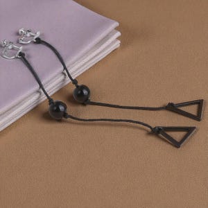 Puede incluir: Pendientes colgantes con cuentas negras y dijes triangulares. Los pendientes tienen cierres de clip plateados y están suspendidos de cordones negros. Descansan sobre tela lavanda y una superficie marrón claro.