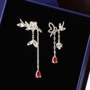 以下が含まれることがあります： シルバーカラーのドロップピアス。片方は花のモチーフ、もう片方は蝶のデザイン。どちらもチェーンの先に涙型の赤い宝石が付いています。ピアスは濃い青色の背景に配置されています。