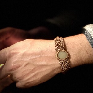 May include: A woven macrame bracelet in shades of brown, featuring a central oval-shaped, light-colored stone. The bracelet is worn on a wrist, showcasing the intricate knotting pattern and the neutral tones of the jewelry.